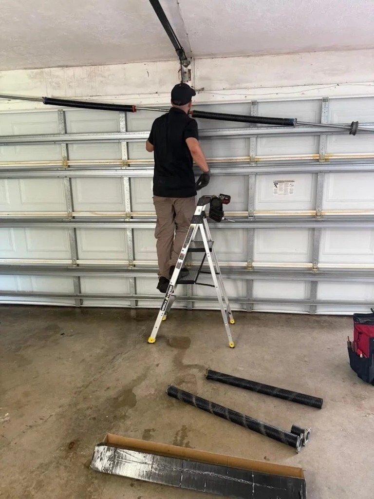 Technician on a stepladder at the top of a garage door, with spring tubes laid on the floor