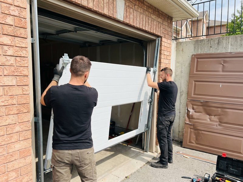 Two technicians lifting a white garage door panel into place at a brick-front garage