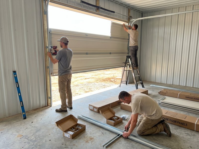 Two installers assembling a new steel garage door in the opening, with track pieces and boxes on the floor